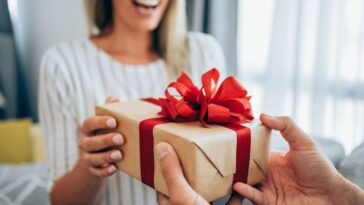 Cropped shot of a loving husband giving his wife a gift. Boyfriend surprises his beautiful girlfriend with present while she is sitting on the sofa in the living room at home. Focus is on the gift.