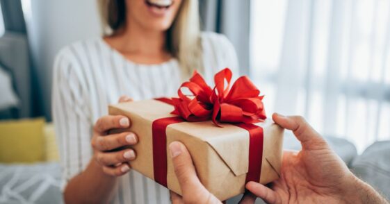 Cropped shot of a loving husband giving his wife a gift. Boyfriend surprises his beautiful girlfriend with present while she is sitting on the sofa in the living room at home. Focus is on the gift.