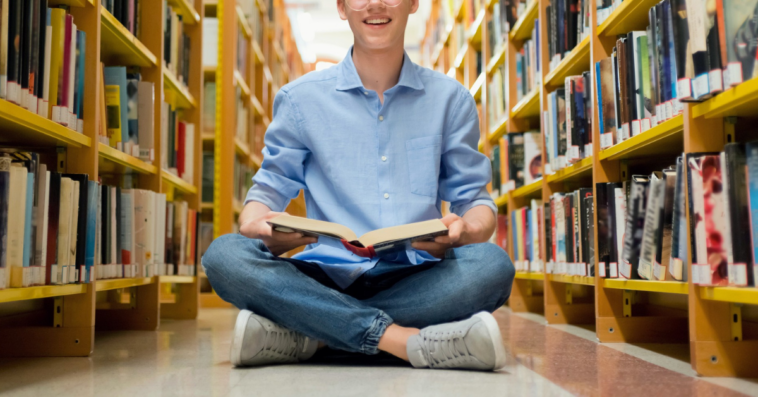 teen boy sitting on library floor looking at a book