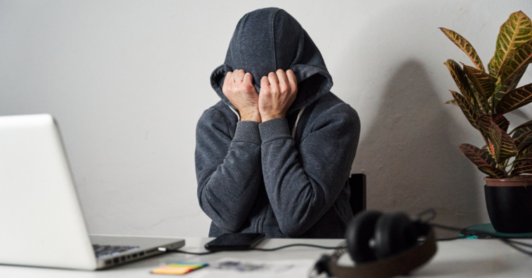 person seated at desk with laptop and hood pulled over head and face