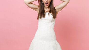 Portrait of an irritated, dissatisfied bride in a white wedding dress standing, screaming and clinging to head; isolated on pink pastel background.