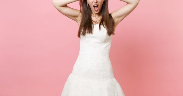 Portrait of an irritated, dissatisfied bride in a white wedding dress standing, screaming and clinging to head; isolated on pink pastel background.