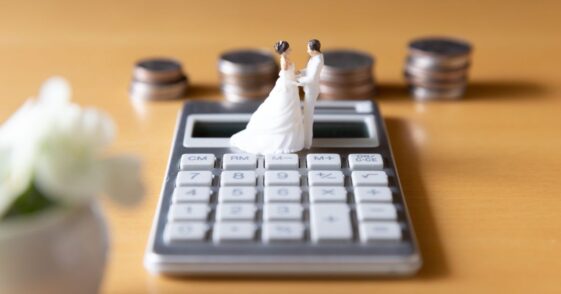 A plastic figure of a married couple sits on top of a calculator. Stacks of coins sit behind them against a yellow background.