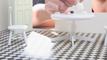 A little girl reaching into a dollhouse and playing with a doll table and chairs.