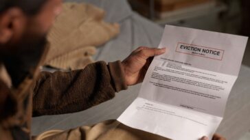 A bearded man holding an eviction notice while sitting on a couch and reading it in a dimly lit room without natural light coming in through the window behind.