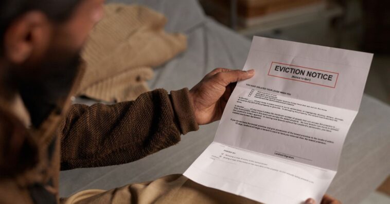 A bearded man holding an eviction notice while sitting on a couch and reading it in a dimly lit room without natural light coming in through the window behind.