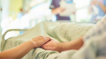 A hospital visitor's hand holds a patient's hand in the bed of a hospital ward. In the blurred background a young nurse is chatting to the ward sister about the patient's care.