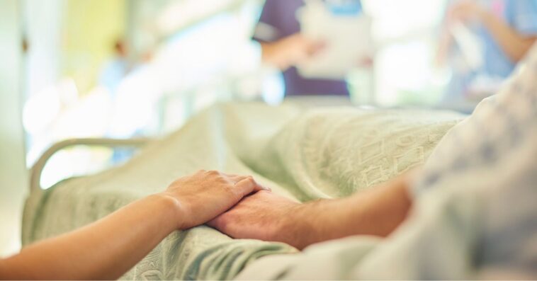 A hospital visitor's hand holds a patient's hand in the bed of a hospital ward. In the blurred background a young nurse is chatting to the ward sister about the patient's care.