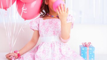 A young girl all dressed up celebrating her birthday, blowing a pink balloon sitting on white sofa in a living room.