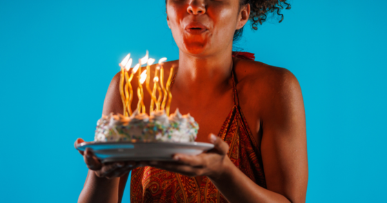 A woman holding a cake with birthday candles in it, blowing them out.