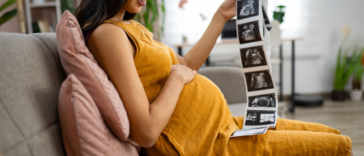 A pregnant woman resting her hand on her belly and holding sonogram pictures.