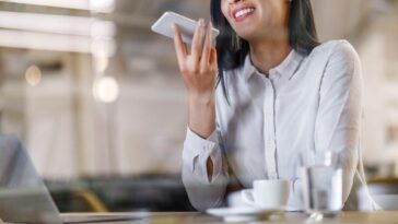 Happy businesswoman talking to virtual assistant on her phone while working in the office. The view is through glass.