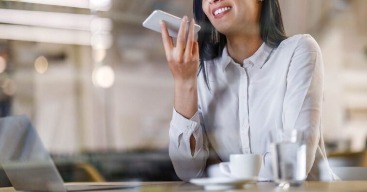 Happy businesswoman talking to virtual assistant on her phone while working in the office. The view is through glass.