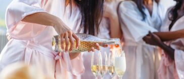 A cheerful bride in a robe pours champagne into glasses at a bachelorette party with friends in pastel robes, celebrating outdoors.