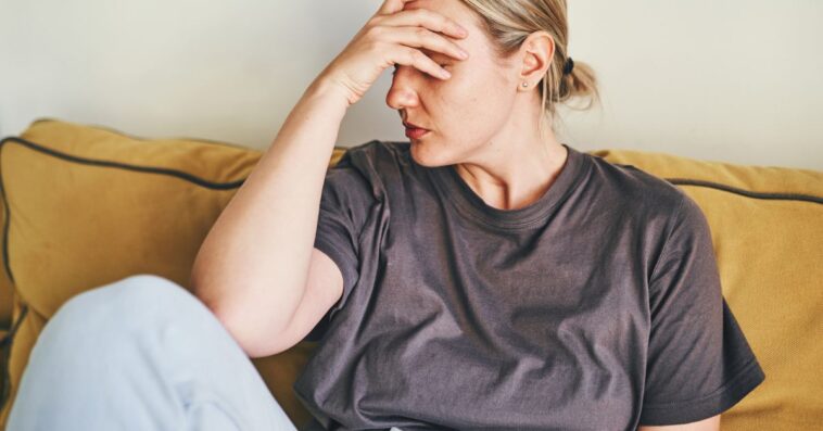 A young woman is sitting at home on a yellow sofa with her head in her hands, sad or showing a headache or frustration and depression.