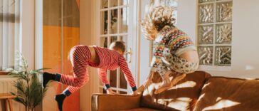 Two cute kids wearing pyjamas bounce on a brown leather couch in a sunny domestic room.