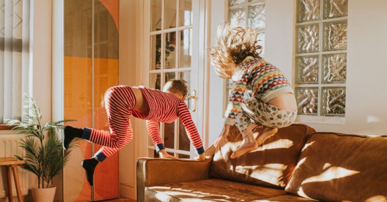 Two cute kids wearing pyjamas bounce on a brown leather couch in a sunny domestic room.