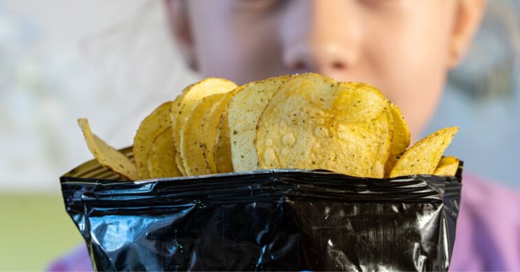 A teenage girl looks with surprise and joy at an open pack of chips.