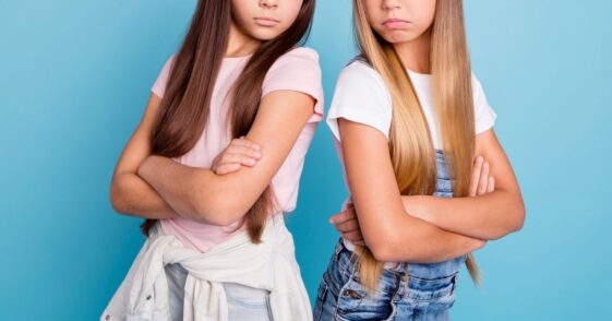 Two frowning adolescent girls stand back to back with their arms folded.