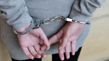 Back shot of a woman dressed in a gray sweatshirt and black pants stands with her hands cuffed behind her back.