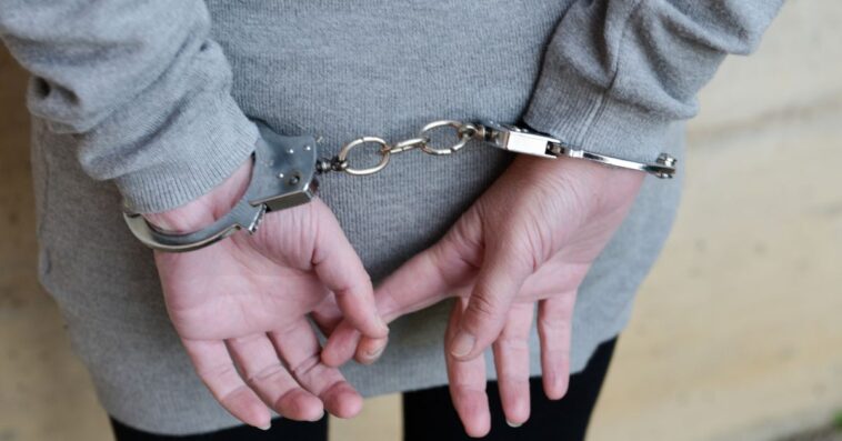 Back shot of a woman dressed in a gray sweatshirt and black pants stands with her hands cuffed behind her back.