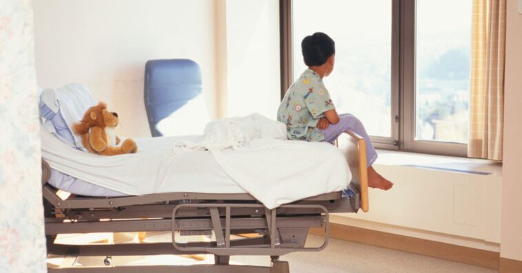 Backshot of a young boy (6-7) sitting on the edge of a hospital bed while gazing out the window.