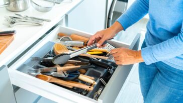 Unrecognizable woman organizing a messy kitchen drawer with a lot of kitchen utensils.