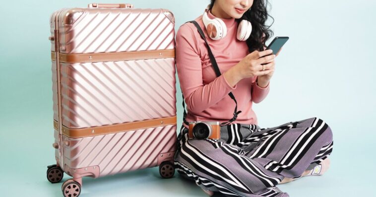 Portrait of a young, pretty girl, sitting next to a large, aluminium suitcase. She is texting and wears large headphones around her neck.