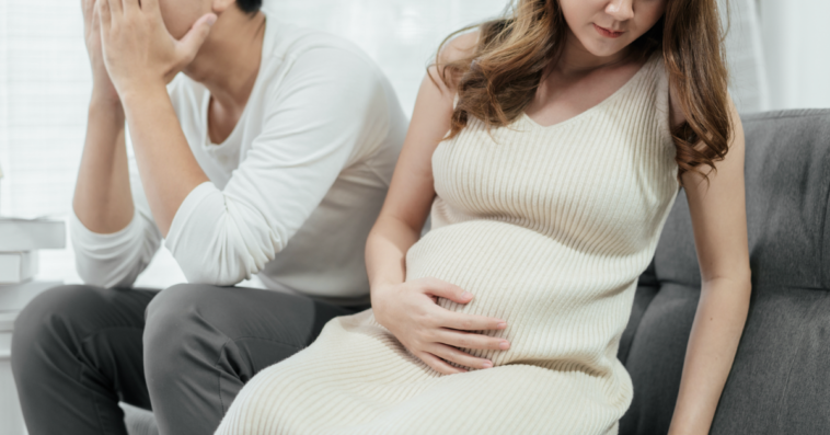 A man sitting on the sofa with his head in his hands sitting next to a pregnant woman.