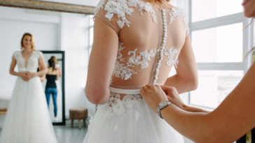 A woman buttoning up a woman in a wedding dress from behind.
