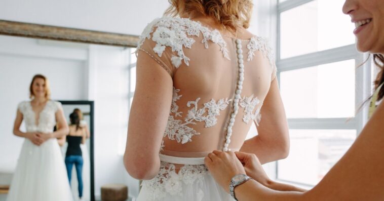 A bride with a dressmaker are making final touch on tailor made gown in bridal clothing shop.