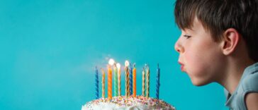 Boy blowing out candles on a birthday cake against blue background.