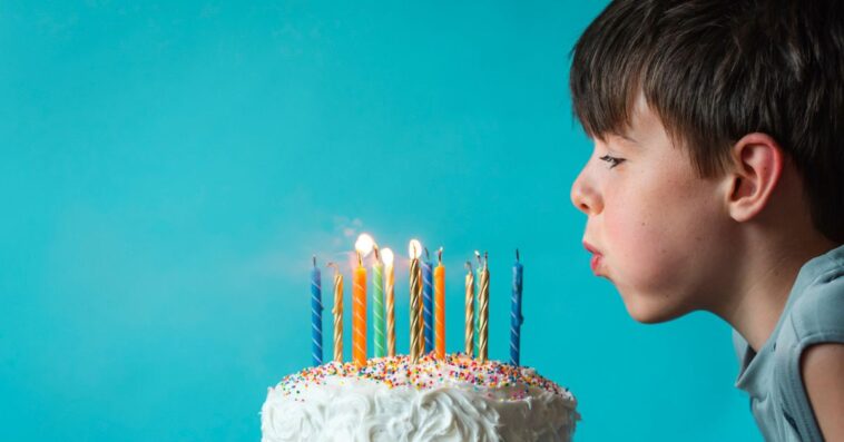 Boy blowing out candles on a birthday cake against blue background.