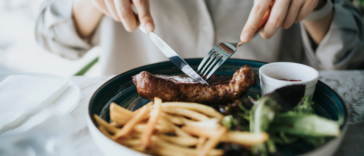 A woman cutting into a steak with french fries and salad greens sharing the plate.