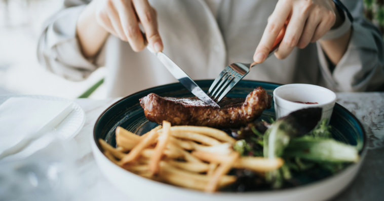 A woman cutting into a steak with french fries and salad greens sharing the plate.