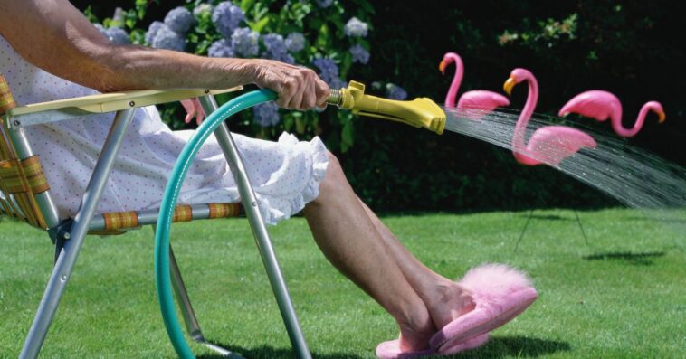 Senior woman in lawn chair, watering grass, low section, side view. Three pink flamingos sit in the background.