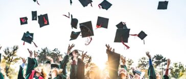Large group of happy college students celebrating their graduation day outdoors while throwing their green caps up in the air.