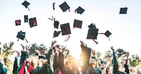 Large group of happy college students celebrating their graduation day outdoors while throwing their green caps up in the air.