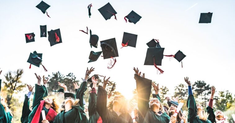 Large group of happy college students celebrating their graduation day outdoors while throwing their green caps up in the air.