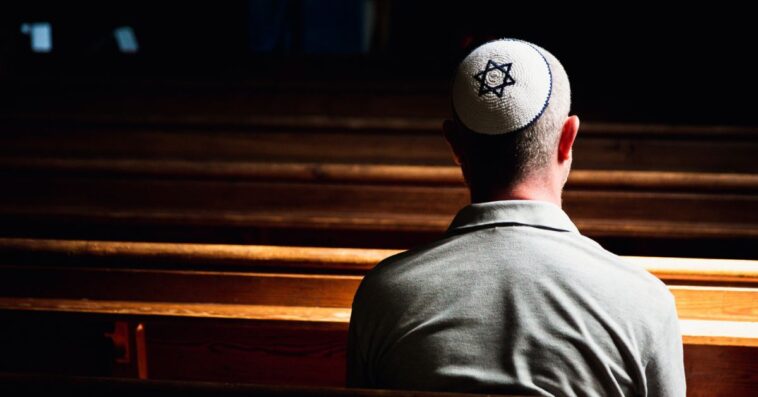 Close up, back-shot image depicting a young caucasian Jewish adult man in his 30s inside a synagogue. He has his head bowed in prayer and he is wearing the traditional Jewish skull cap - otherwise known as a kippah or yarmulke - on his head. The man has a beard and the background of the synagogue is blurred out of focus. Horizontal color image with copy space.