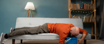 A man lying on his stomach on the sofa and throwing up into a bucket.
