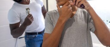 Medium point-of-view shot of a teenage boy and his father standing in the bathroom of their home in Sunderland, North East England. The boy is using an electric razor to trim his facial hair, he is distorting his face as he shaves one cheek. His father is smiling at him in the background.