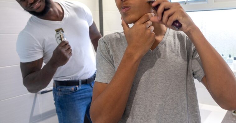 Medium point-of-view shot of a teenage boy and his father standing in the bathroom of their home in Sunderland, North East England. The boy is using an electric razor to trim his facial hair, he is distorting his face as he shaves one cheek. His father is smiling at him in the background.