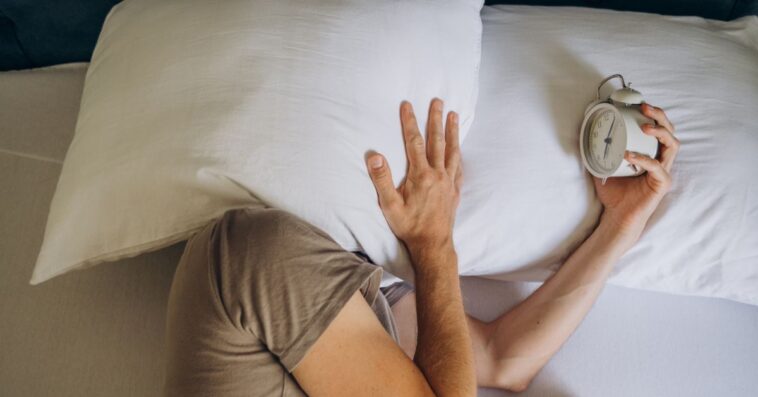 A man laying in bed holds his pillow over his face. He holds out an alarm clock.
