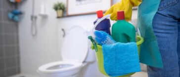 A cleaning woman is standing in the bathroom holding a blue bucket full of chemicals and facilities. The toilet is in the corner.