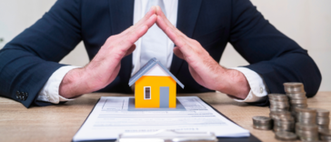 A man in a suit sitting at a desk with his fingers touching over a contract and a miniature house.