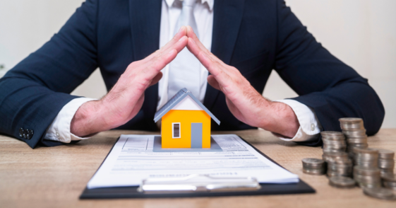 A man in a suit sitting at a desk with his fingers touching over a contract and a miniature house.
