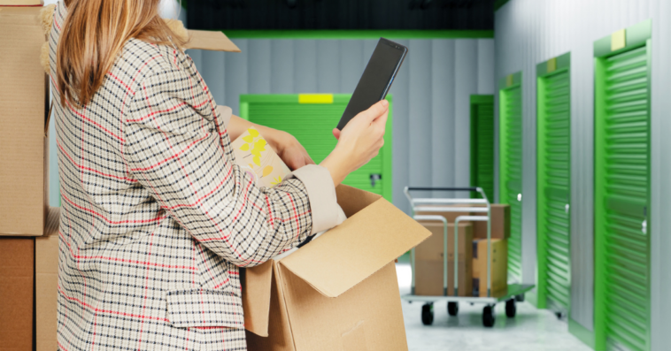 A woman on her phone holding a box in a storage facility.