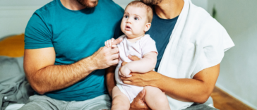 gay couple hold their baby while seated on a bed