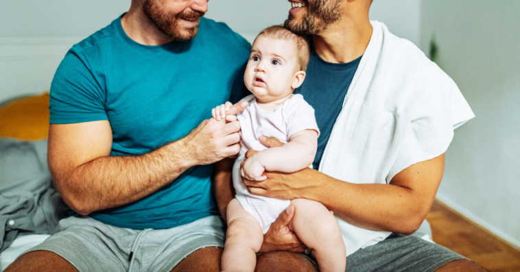 gay couple hold their baby while seated on a bed